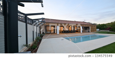 Luxurious modern villa at twilight, featuring a serene swimming pool reflecting warm interior lights. An inviting wooden deck hosts an outdoor dining area, complemented by a stylish hanging chair Luxurious modern villa at twilight, featuring a serene swimming pool reflecting warm interior lights. An inviting wooden deck hosts an outdoor dining area, complemented by a stylish hanging chair 134575549