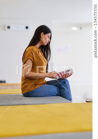 Woman reading a magazine while sitting on a bench in a bright, modern indoor space 134575550