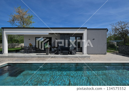 Modern modular house backyard featuring a rectangular swimming pool, large patio with wooden sun loungers, and umbrellas against a blue sky. Contemporary prefabricated home design 134575552