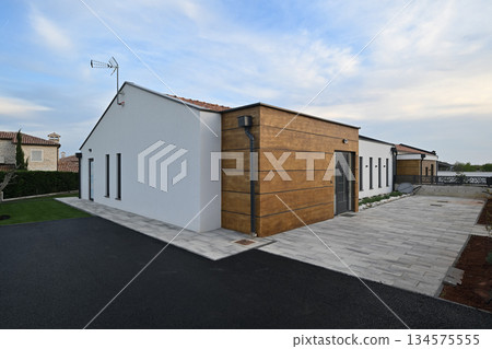 Modern house entrance with a stylish dark door, striking wooden facade, and paved driveway. Sunlight flares through the dark metal gate, adding a dynamic touch to this contemporary home's exterior Modern house entrance with a stylish dark door, striking wooden facade, and paved driveway. Sunlight flares through the dark metal gate, adding a dynamic touch to this contemporary home's exterior 134575555