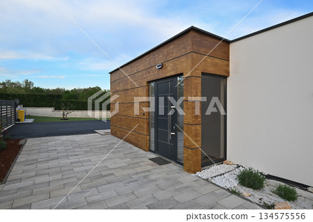 Modern house entrance with a stylish dark door, striking wooden facade, and paved driveway. Sunlight flares through the dark metal gate, adding a dynamic touch to this contemporary home's exterior 134575556