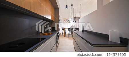 Modern open-plan living and dining area with an integrated kitchen, featuring a comfortable sofa, stylish armchairs, and a bar-height dining table. Wooden floors and ceiling beams enhance this Modern open-plan living and dining area with an integrated kitchen, featuring a comfortable sofa, stylish armchairs, and a bar-height dining table. Wooden floors and ceiling beams enhance this 134575595