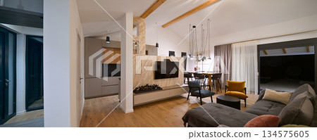 Modern open-plan living and dining area with an integrated kitchen, featuring a comfortable sofa, stylish armchairs, and a bar-height dining table. Wooden floors and ceiling beams enhance this Modern open-plan living and dining area with an integrated kitchen, featuring a comfortable sofa, stylish armchairs, and a bar-height dining table. Wooden floors and ceiling beams enhance this 134575605