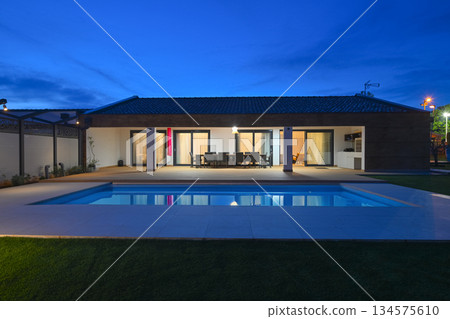 Luxurious modern villa at twilight, featuring a serene swimming pool reflecting warm interior lights. An inviting wooden deck hosts an outdoor dining area, complemented by a stylish hanging chair Luxurious modern villa at twilight, featuring a serene swimming pool reflecting warm interior lights. An inviting wooden deck hosts an outdoor dining area, complemented by a stylish hanging chair 134575610