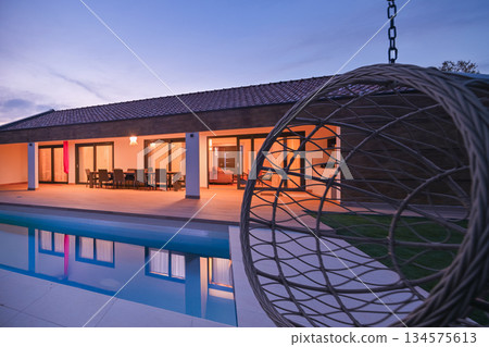 Luxurious modern villa at twilight, featuring a serene swimming pool reflecting warm interior lights. An inviting wooden deck hosts an outdoor dining area, complemented by a stylish hanging chair Luxurious modern villa at twilight, featuring a serene swimming pool reflecting warm interior lights. An inviting wooden deck hosts an outdoor dining area, complemented by a stylish hanging chair 134575613