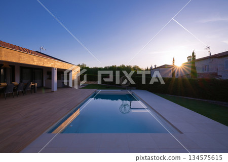 Modern villa with a stunning swimming pool and spacious wooden deck, perfect for outdoor dining. Capturing the golden hour with a beautiful sunset and clear sky, showcasing luxury living and serene Modern villa with a stunning swimming pool and spacious wooden deck, perfect for outdoor dining. Capturing the golden hour with a beautiful sunset and clear sky, showcasing luxury living and serene 134575615