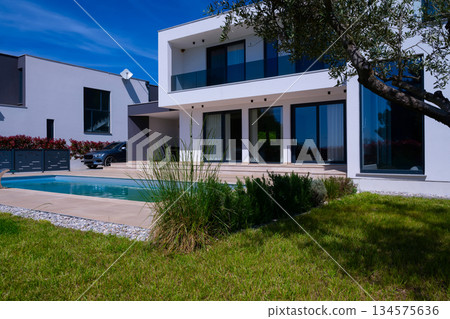 Modern luxury villa with swimming pool and clean minimalist architecture under a blue sky. Contemporary residential exterior featuring large windows, terrace, and elegant outdoor living space Modern luxury villa with swimming pool and clean minimalist architecture under a blue sky. Contemporary residential exterior featuring large windows, terrace, and elegant outdoor living space 134575636