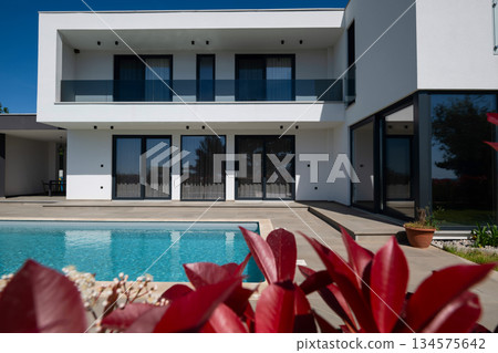 Modern luxury villa with swimming pool and clean minimalist architecture under a blue sky. Contemporary residential exterior featuring large windows, terrace, and elegant outdoor living space 134575642