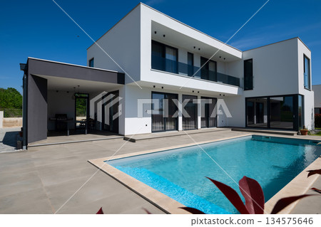 Modern luxury villa with swimming pool and clean minimalist architecture under a blue sky. Contemporary residential exterior featuring large windows, terrace, and elegant outdoor living space Modern luxury villa with swimming pool and clean minimalist architecture under a blue sky. Contemporary residential exterior featuring large windows, terrace, and elegant outdoor living space 134575646