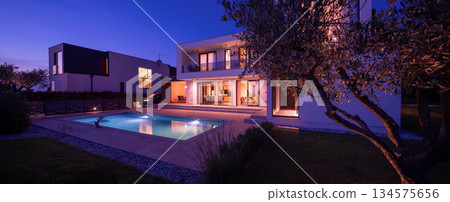 Modern luxury villa with a luminous swimming pool at twilight. Contemporary architecture, sleek design, and inviting interior lights create a serene and elegant evening ambiance, ideal for upscale 134575656