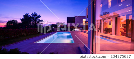Modern luxury villa with a luminous swimming pool at twilight. Contemporary architecture, sleek design, and inviting interior lights create a serene and elegant evening ambiance, ideal for upscale 134575657