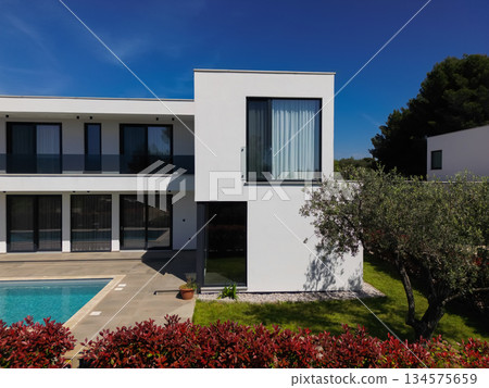 Modern luxury villa with swimming pool and clean minimalist architecture under a blue sky. Contemporary residential exterior featuring large windows, terrace, and elegant outdoor living space Modern luxury villa with swimming pool and clean minimalist architecture under a blue sky. Contemporary residential exterior featuring large windows, terrace, and elegant outdoor living space 134575659