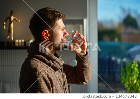 Man drinking water from glass in kitchen interior with daylight Man drinking water from glass in kitchen interior with daylight 134578348