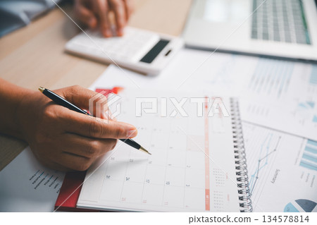 Office worker marking calendar while calculating financial data, illustrating schedule planning, time management, business tasks, productivity, professional workflow in modern workplace environment. 134578814