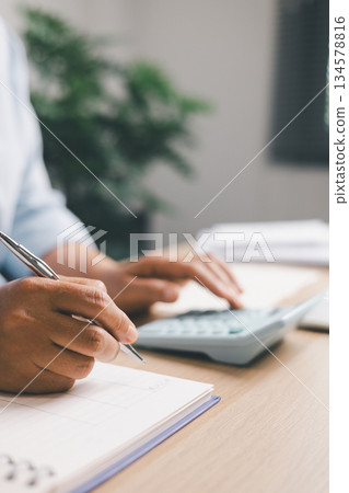Close-up of office worker writing financial figures in notebook while using a calculator, illustrating budgeting, accounting tasks, expense tracking, professional financial management in workplace. 134578816