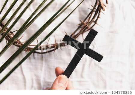 Hand holding wooden cross over white fabric and green palm leaves, symbolizing Christian faith, Palm Sunday, worship and spiritual devotion, minimal religious concept with natural light background. 134578836