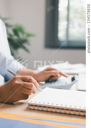 Close-up of office worker writing financial records in notebook while holding calculator, illustrating accounting tasks, expense tracking, budgeting and professional financial management in workplace 134578838