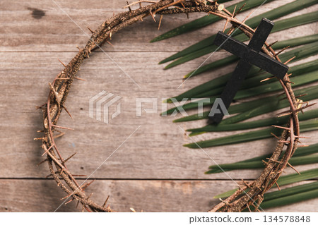 Black wooden cross with crown of thorns, palm leaves on rustic wooden table, symbolizing Christian faith, sacrifice, Palm Sunday, Easter reflection, religious background for worship. Black wooden cross with crown of thorns, palm leaves on rustic wooden table, symbolizing Christian faith, sacrifice, Palm Sunday, Easter reflection, religious background for worship. 134578848