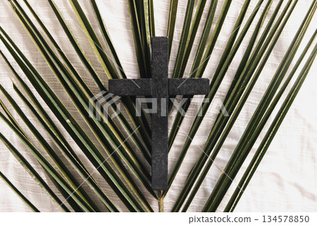 Wooden cross on green palm leaves with crown of thorns over white fabric background, symbolizing Christian faith, Palm Sunday, sacrifice, Easter reflection, minimal religious concept with copy space. Wooden cross on green palm leaves with crown of thorns over white fabric background, symbolizing Christian faith, Palm Sunday, sacrifice, Easter reflection, minimal religious concept with copy space. 134578850