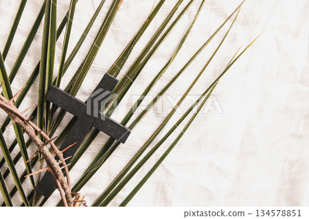 Wooden cross on green palm leaves with crown of thorns over white fabric background, symbolizing Christian faith, Palm Sunday, sacrifice, Easter reflection, minimal religious concept with copy space. 134578851