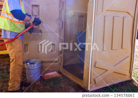 Worker sprays water inside portable toilet gray bin are nearby on ground. Worker sprays water inside portable toilet gray bin are nearby on ground. 134578861