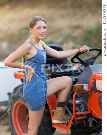 Girl in summer sundress near tractor Girl in summer sundress near tractor 134579571