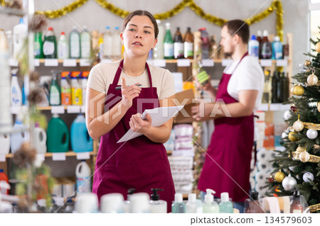 Attentive saleswoman restock of goods, checking papers, male assistant laying out goods in background in small household store decorated for New Year celebration 134579603