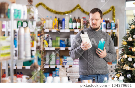 Young man looking shampoo or shower gel, standing near shelves in beauty store decorated for the New Year holiday 134579691