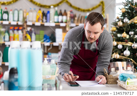 Attentive male seller taking shopping orders, writing down in hardware department of supermarket decorated for New Year celebration Attentive male seller taking shopping orders, writing down in hardware department of supermarket decorated for New Year celebration 134579717