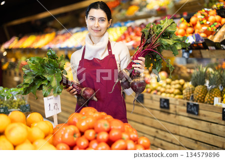 Woman staff with beetroot in hands working in greengrocery Woman staff with beetroot in hands working in greengrocery 134579896