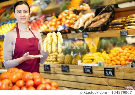Positive female seller offers to buy ripe tasty bananas in grocery department of supermarket 134579967