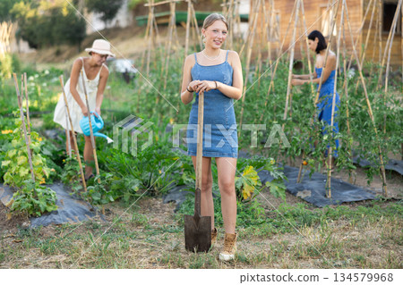 Girl near bed of greenhouse plants. Two garden plot workers are busy in background Girl near bed of greenhouse plants. Two garden plot workers are busy in background 134579968