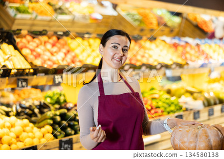 Portrait of positive female grocery supermarket employee near vegetables, fruits and pumpkins Portrait of positive female grocery supermarket employee near vegetables, fruits and pumpkins 134580138