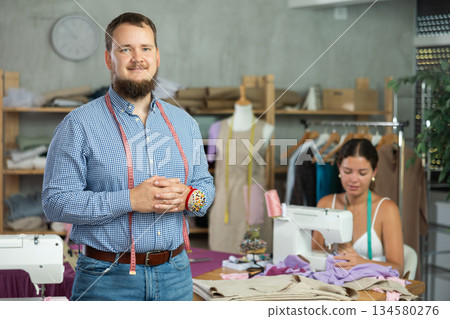 Male dressmaker welcoming clients while young seamstress is sewing behind 134580276