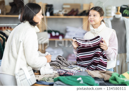 Mother and teen daughter looks at products in store and selects knitted jumper. Mother and teen daughter looks at products in store and selects knitted jumper. 134580311