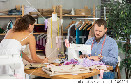Male tailor sewing garment while colleague drafts pattern 134580334