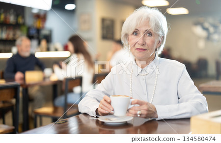 Elderly woman drinking coffee at a table in a cafe Elderly woman drinking coffee at a table in a cafe 134580474