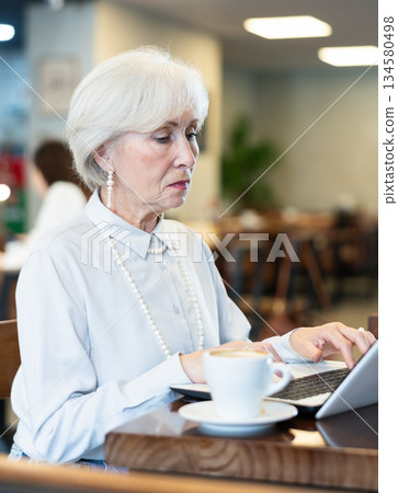 Elderly woman sitting at laptop in cafe Elderly woman sitting at laptop in cafe 134580498