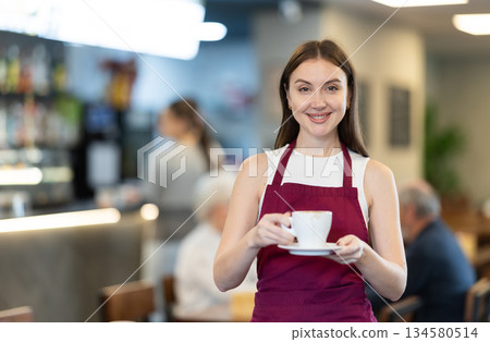 Girl waitress with cup of coffee between rows of tables next to bar counter Girl waitress with cup of coffee between rows of tables next to bar counter 134580514