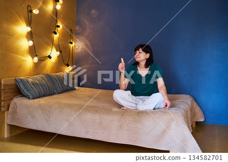Mature Woman sitting on bed in pajamas, enjoying Palo Santo for cozy lifestyle moment Mature Woman sitting on bed in pajamas, enjoying Palo Santo for cozy lifestyle moment 134582701