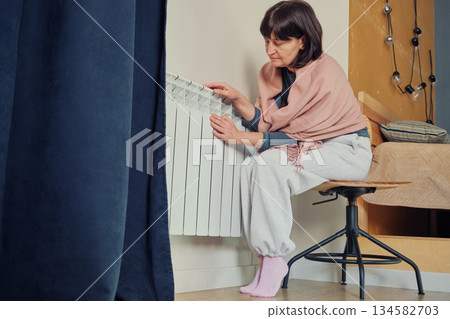 Senior woman at home checking cold radiator during winter, feeling the energy crisis Senior woman at home checking cold radiator during winter, feeling the energy crisis 134582703
