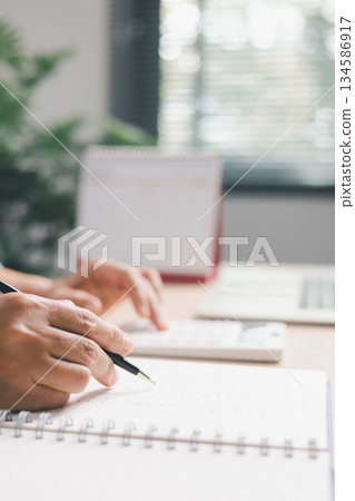 Close-up of office worker calculating numbers, calculator while reviewing financial records in notebook, illustrating accounting tasks, budgeting, expense tracking, professional financial management. 134586917
