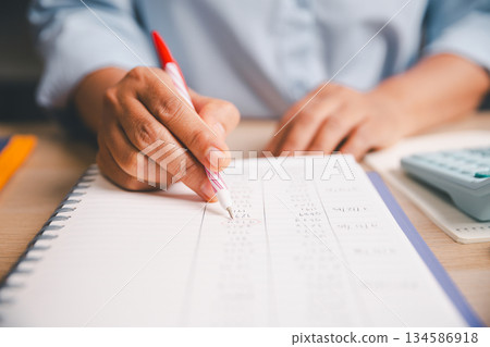 Close-up of office worker marking, writing financial figures in notebook, illustrating accounting tasks, record keeping, expense tracking, budgeting, professional financial management in workplace. 134586918