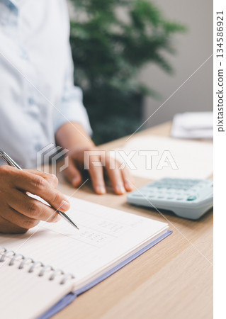 Close-up of office worker writing financial figures in notebook while using a calculator, illustrating budgeting, accounting tasks, expense tracking, professional financial management in workplace. 134586921