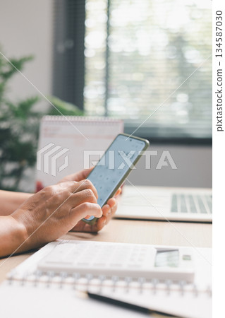 Office worker using smartphone calculator to compute financial numbers at desk, illustrating digital accounting, budgeting, expense tracking, modern workflow and professional financial management. 134587030