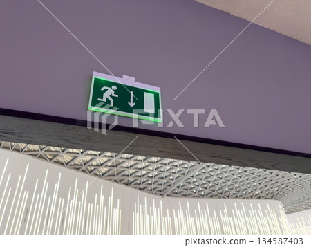 Sign on top of wall with navigational information showing direction to exit building in case of fire. Illuminated sign with running man symbol and arrow pointing toward door for emergency situation Sign on top of wall with navigational information showing direction to exit building in case of fire. Illuminated sign with running man symbol and arrow pointing toward door for emergency situation 134587403