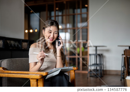 Asian woman talking on phone with tablet on her lap sitting at wooden chair in cafe or coffee shop. Asian woman talking on phone with tablet on her lap sitting at wooden chair in cafe or coffee shop. 134587776