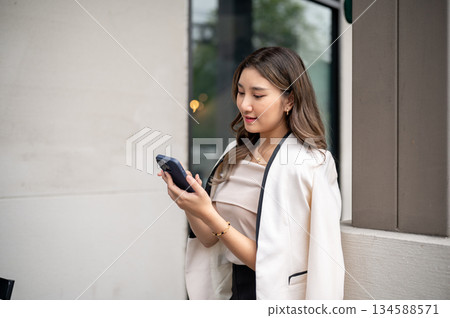 Long hair asian woman smiling as she looking at her smartphone while standing at building's pillar. 134588571