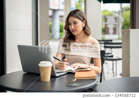 Long hair asian woman looking at laptop while holding pen writing in book with coffee at cafe table. 134588712