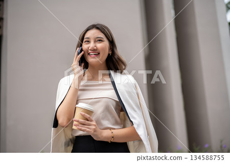 Asian woman smiling and talking on phone while walking around the building with a coffee cup in hand 134588755